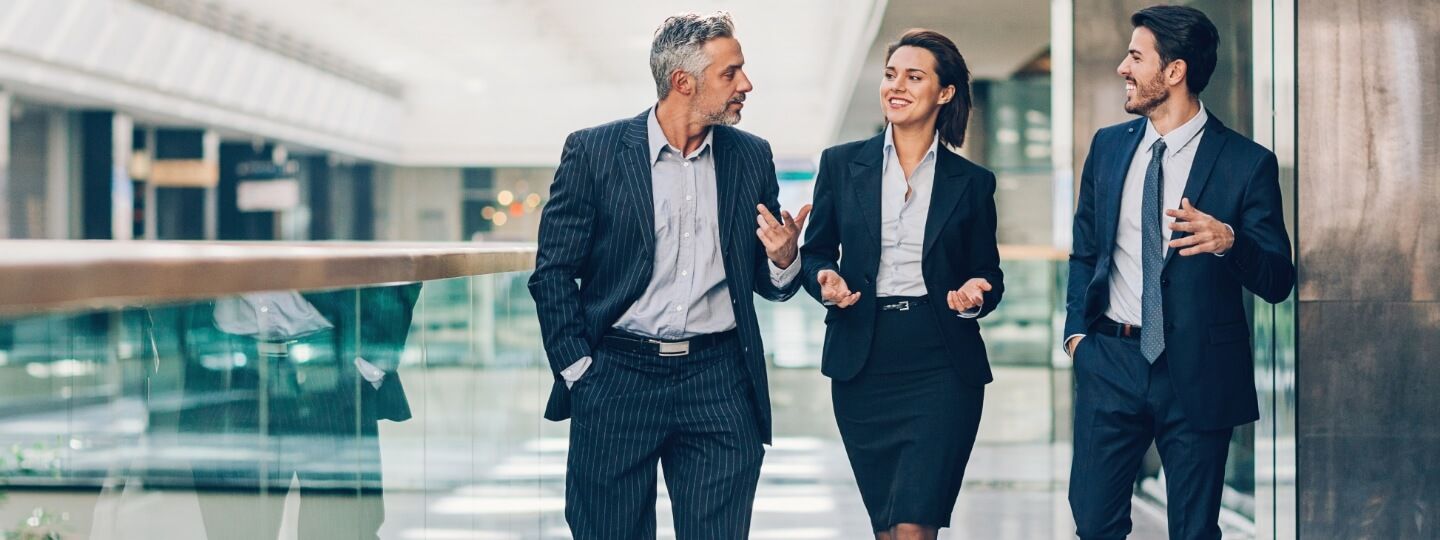 Zwei Männer und eine Frau im Businessoutfit reden während des Gehens in einem Bürogebäude