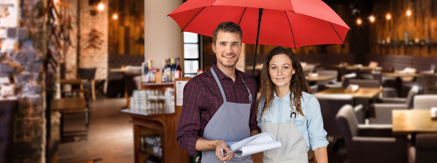 Eine junge Frau und ein junger Mann aus der Gastronomie stehen in ihrem Lokal unter dem roten Schutzschirm.