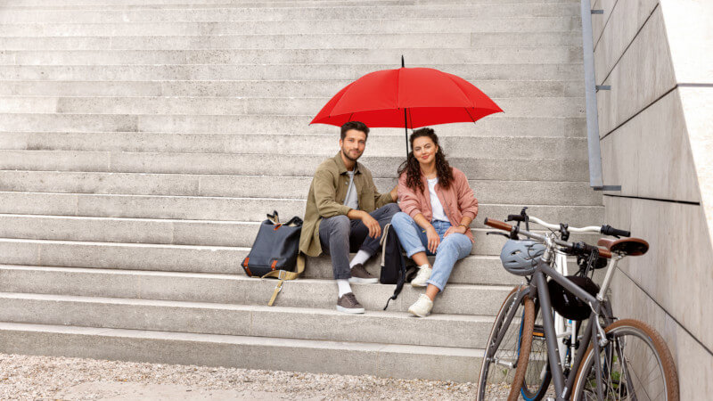 Zwei Personen auf einer Treppe sitzend mit einem roten Schirm über ihnen und einem Fahrrad vor ihnen