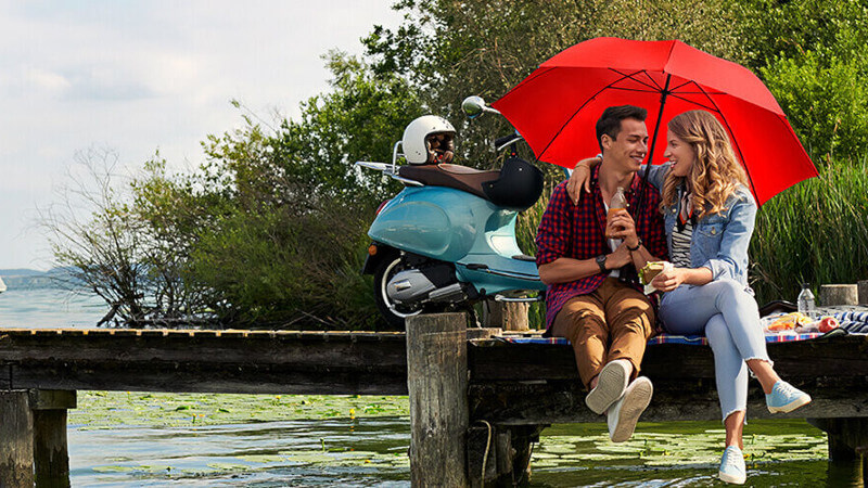 Zwei Personen auf einem Steg mit einem roten Schirm und ihrem Moped im Hintergrund